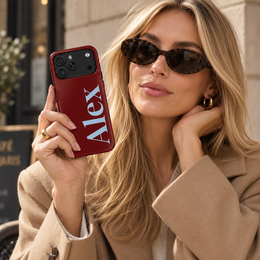 Dark cherry name case printed with the name Alex in powder blue serif lettering, held up by a woman in sunglasses